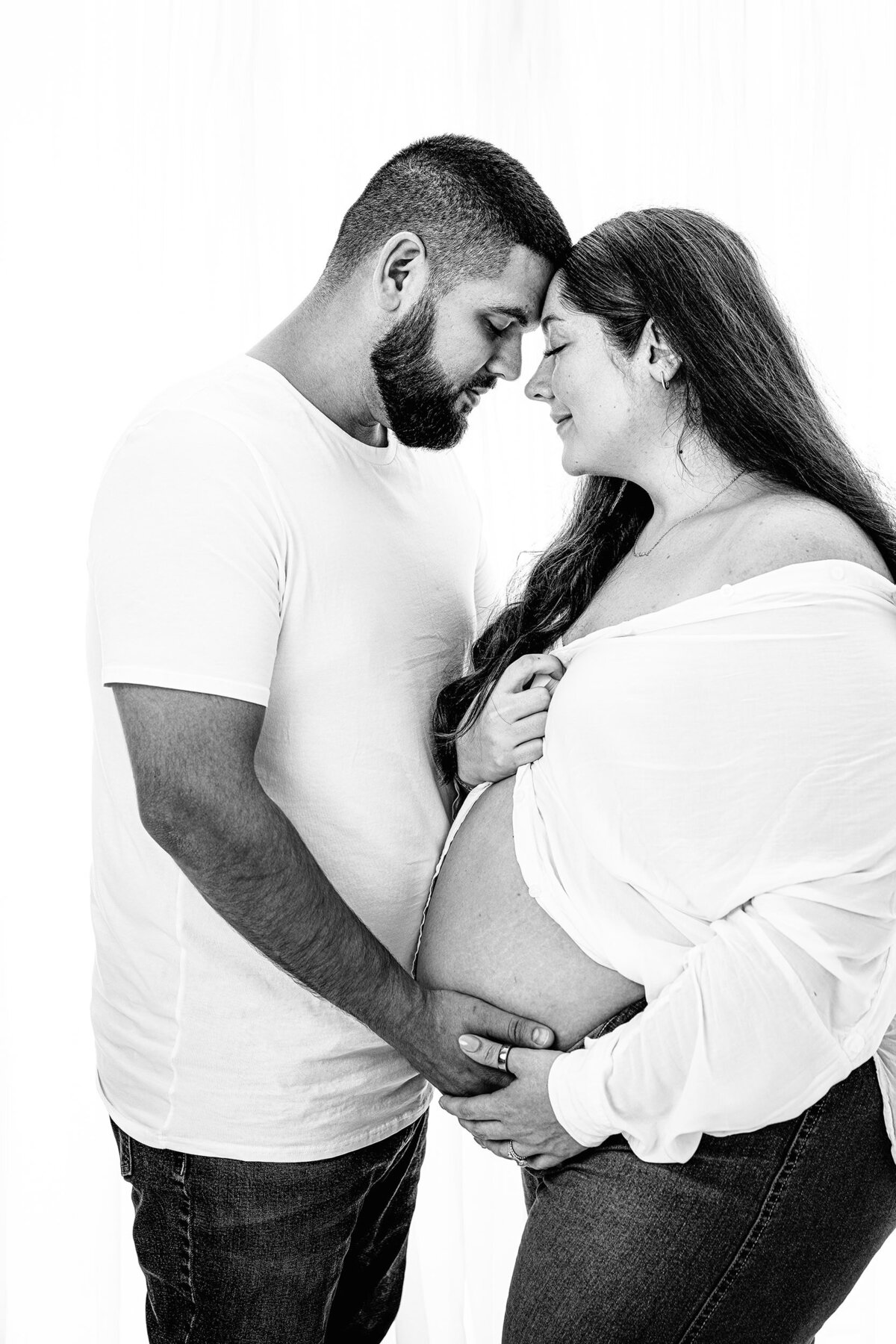 Black and white photo of expectant couple wearing white and heads together in an emotive way