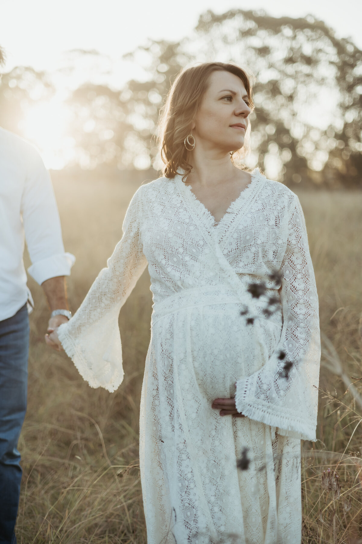 Close up of beautiful pregnant woman holding her bump and leading her husband thrrough a peaceful field during sunset
