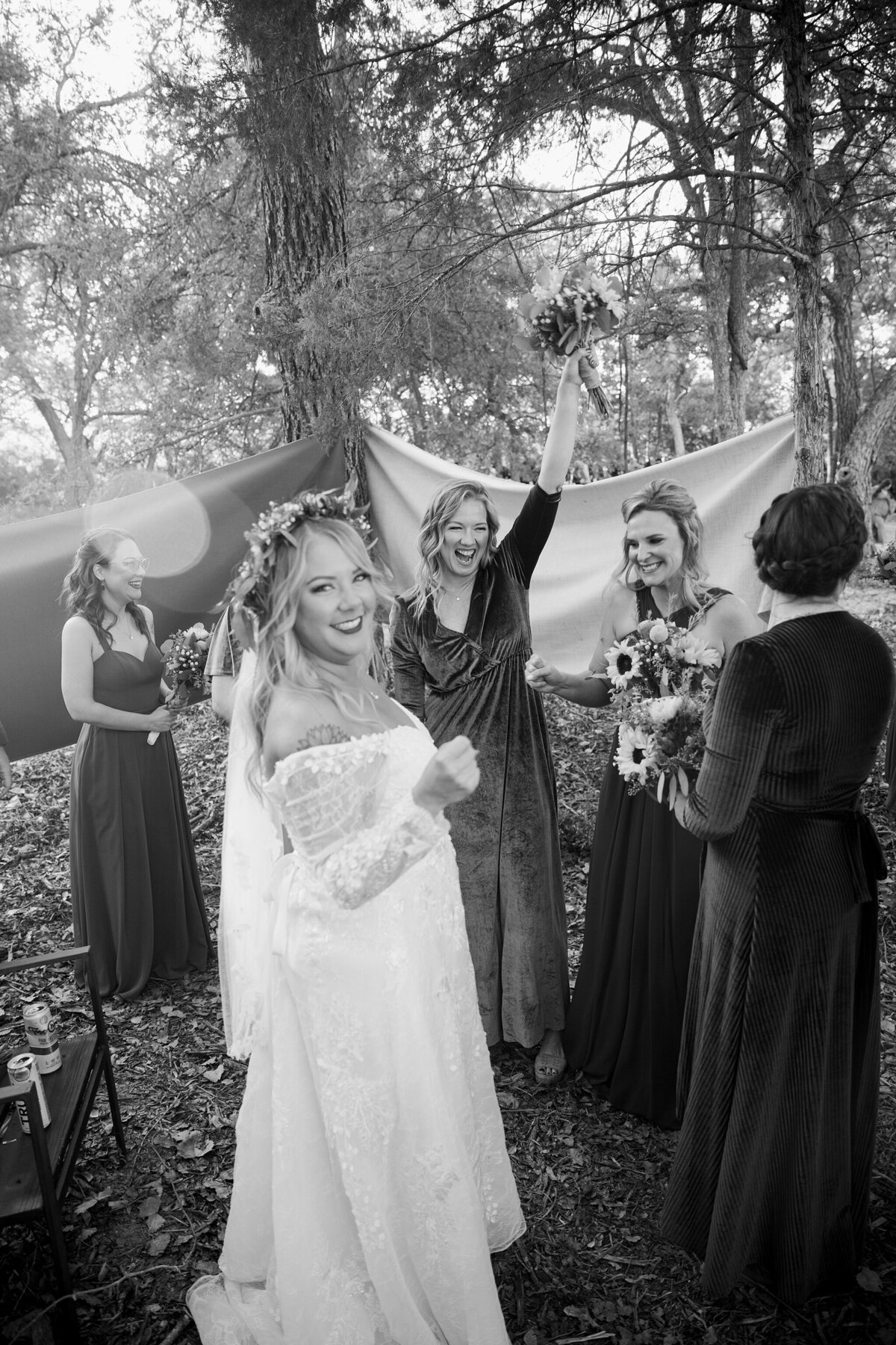 A black and white photo of a bride and her bridesmaids laughing, smiling, and celebrating behind a barrier right after a wedding ceremony in DFW, Texas.  the bride is wearing a long, flowing white dress with a flower crown while her bridesmaids are all wearing dark dresses and holding bouquets.