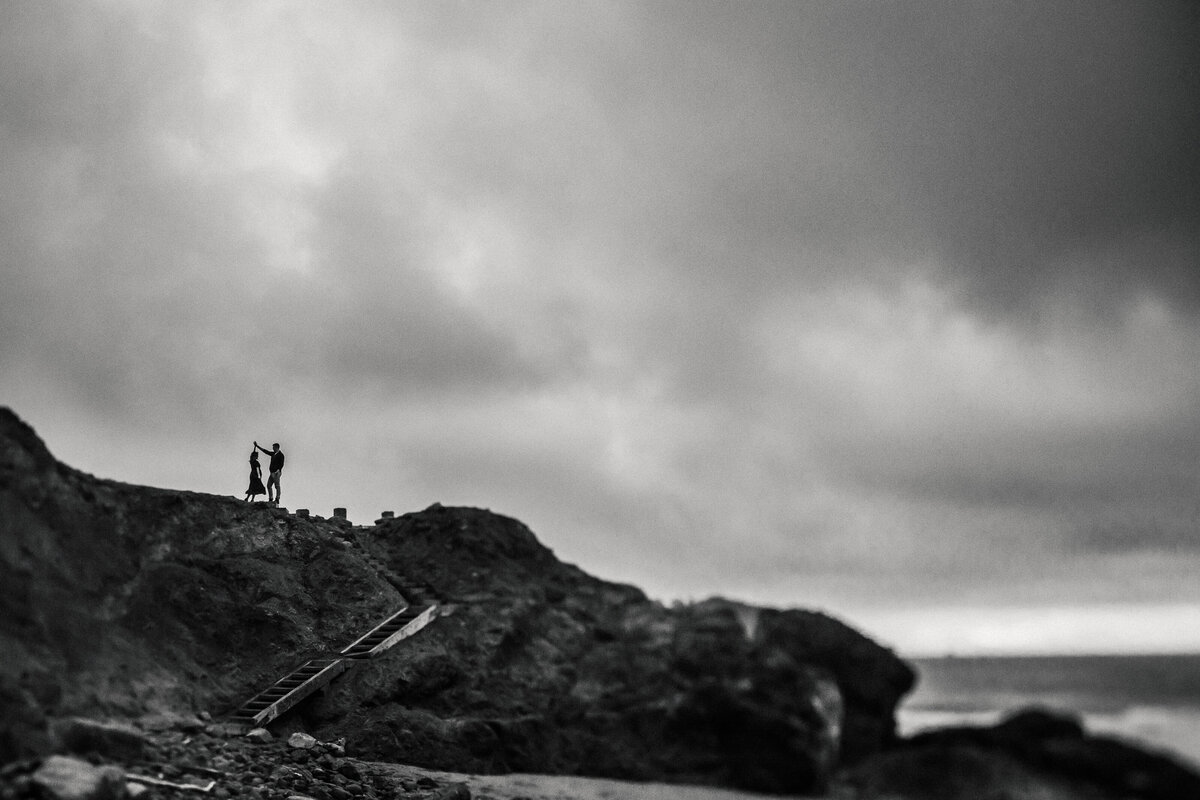 Wide angle portrait of maternity couple dancing on bluffs off california coast. Cloudy sky and photo in black and white