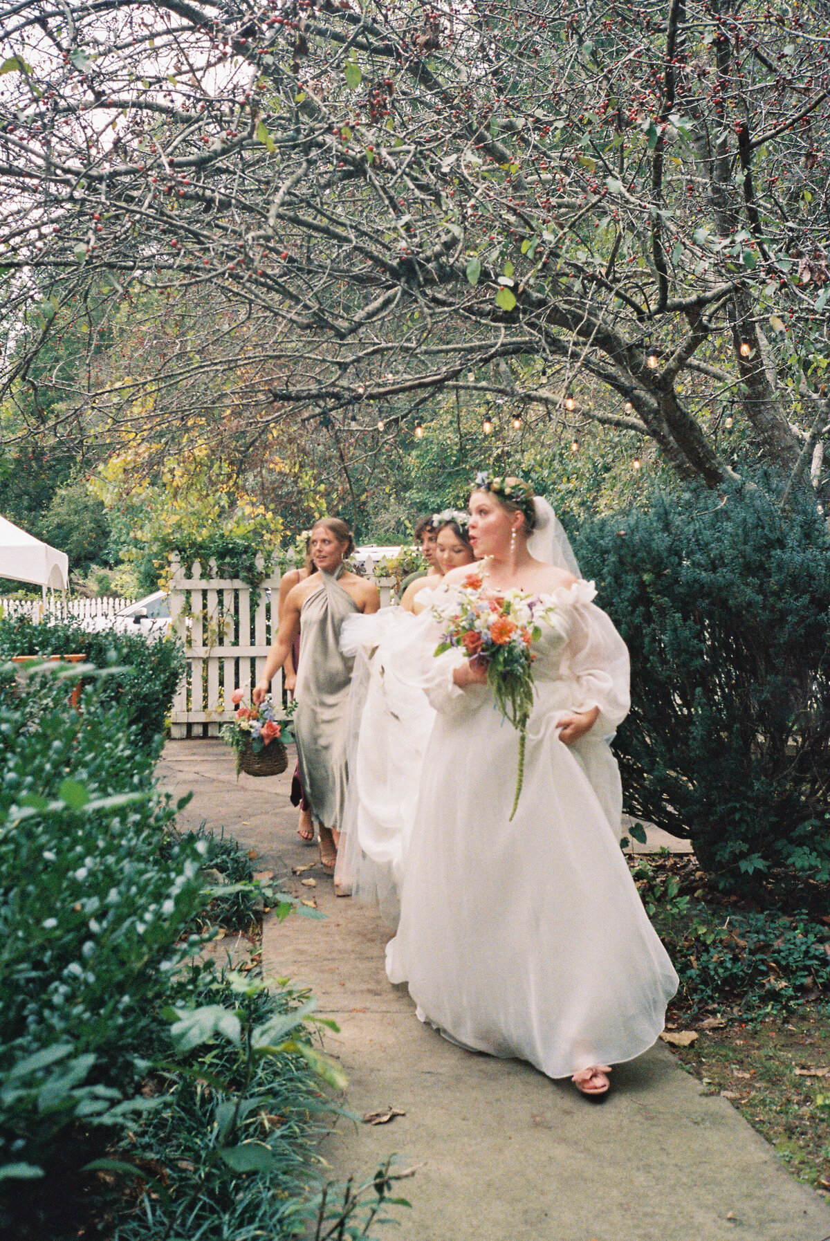 Film photograph of a bride in a romantic off-the-shoulder wedding gown walking down a garden path with her bridesmaids, all holding whimsical garden-style bouquets featuring dahlias, roses, and greenery. Designed by a Northwest Arkansas wedding florist for an autumn outdoor celebration.