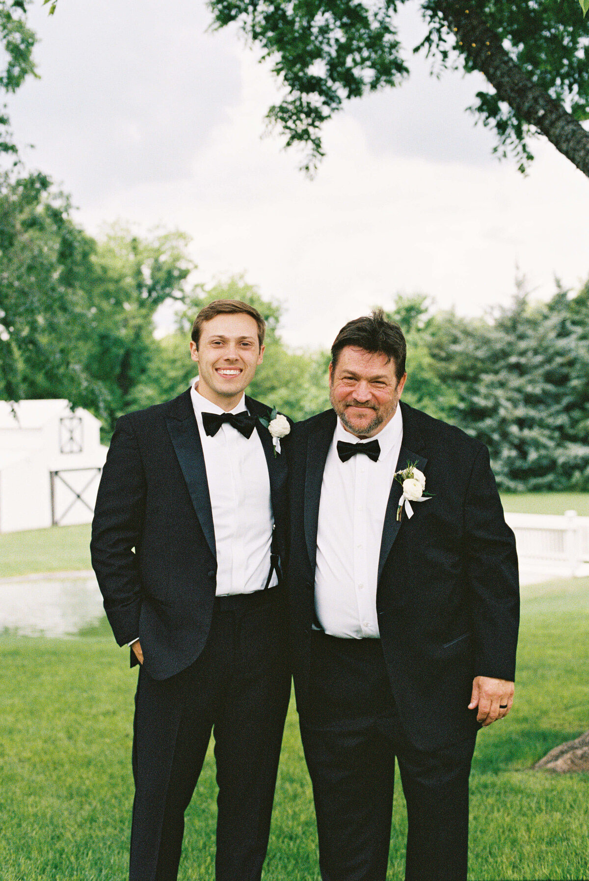 Two men in tuxedos, adorned with white boutonnières, stand smiling on a green lawn. A sunny day with trees and a building in the background.