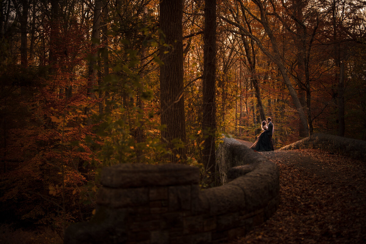 A couple standing on a stone bridge covered in fall colored leaves.