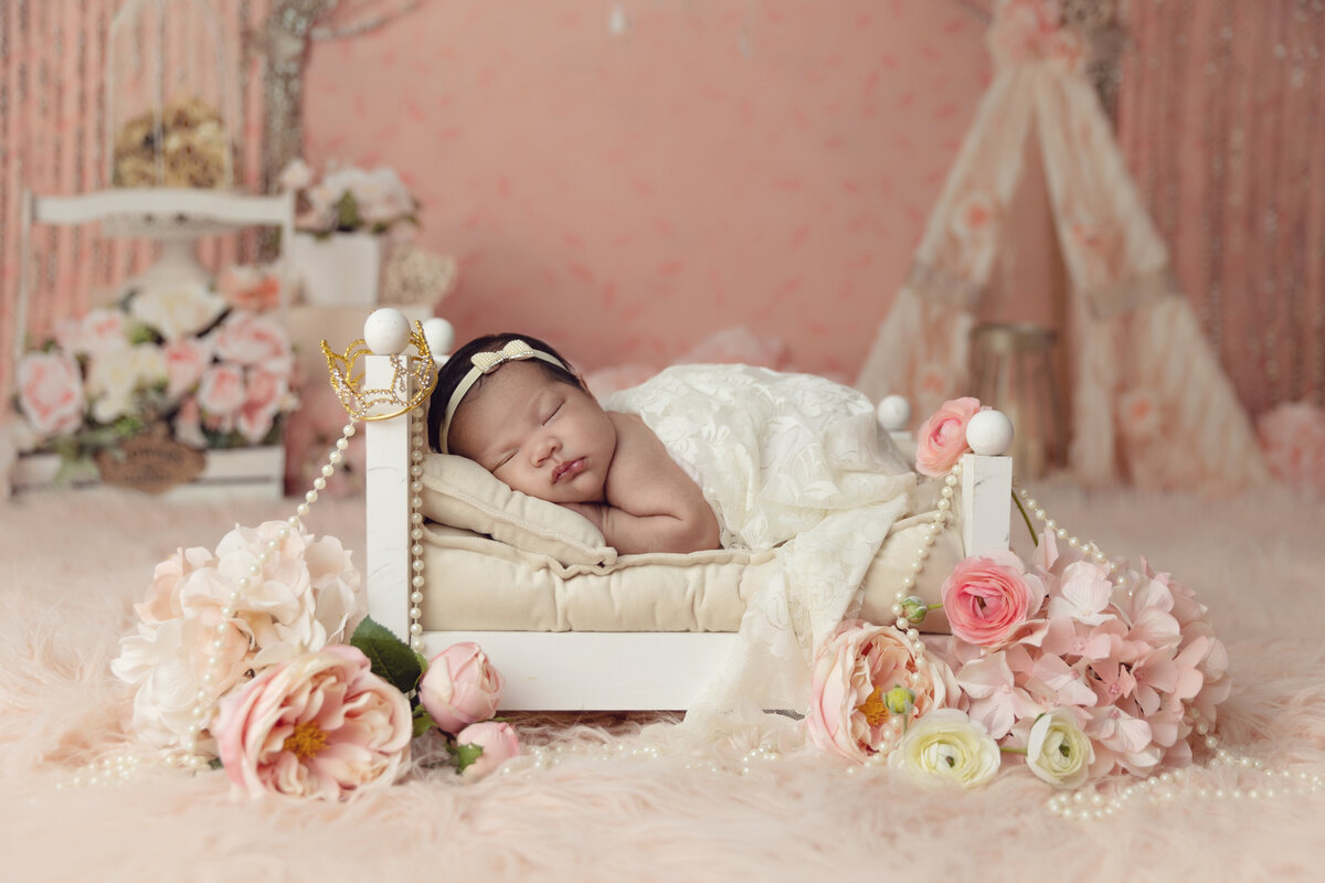 A sleeping newborn baby lays on her stomach in a tiny wooden bed surrounded by flowers