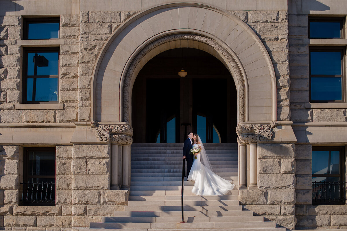 bride and groom stand together on stone steps under arch way