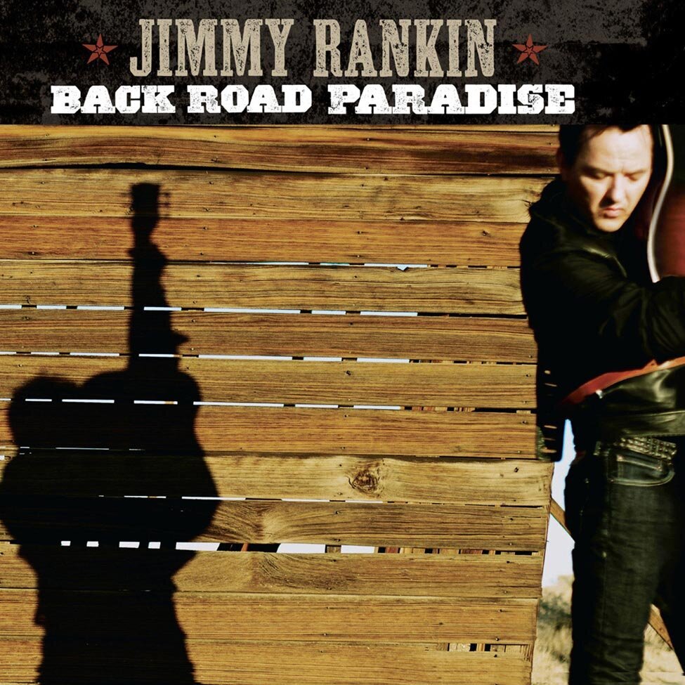 Album Cover Title Back Road Paradise Artist Jimmy Rankin standing in front of barn holding guitar straight up in air shadow on wood wall behind him