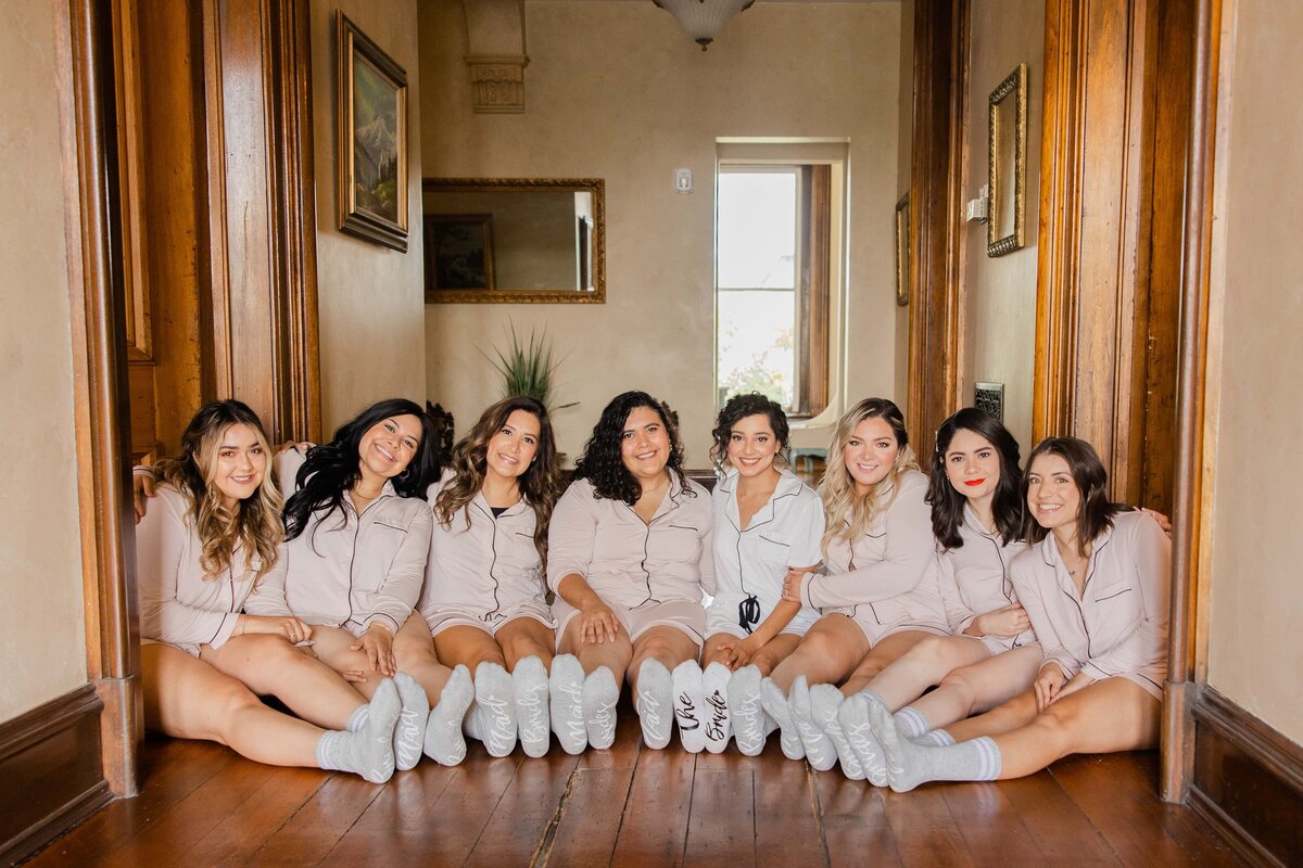 Eight women dressed in matching beige robes and white sneakers sit in a hallway, smiling at the camera, with their feet playfully extended forward during a park farm winery wedding.