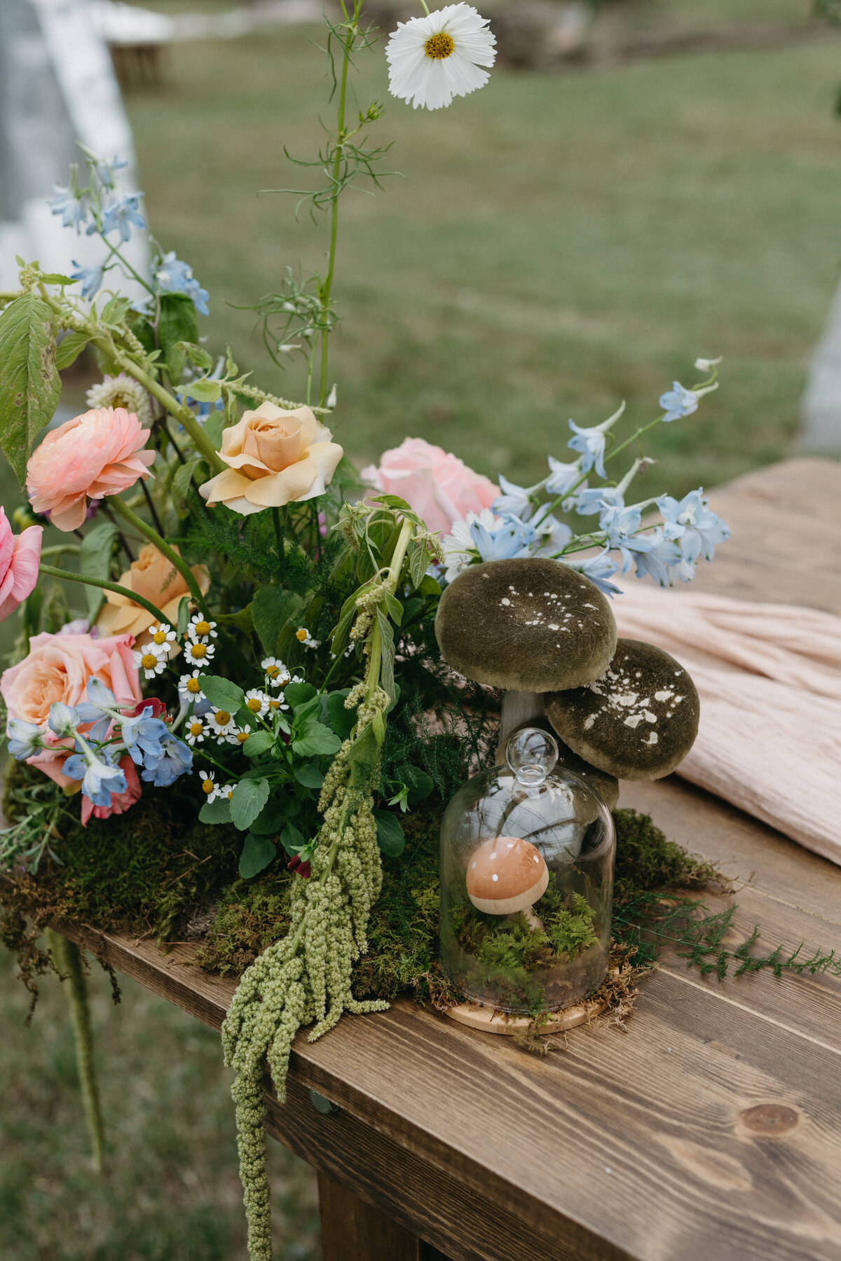 Whimsical garden-style wedding centerpiece with peach ranunculus, yellow roses, chamomile, delphinium, and faux mushrooms on a moss runner for an enchanted forest-inspired reception at The Nest in Northwest Arkansas.