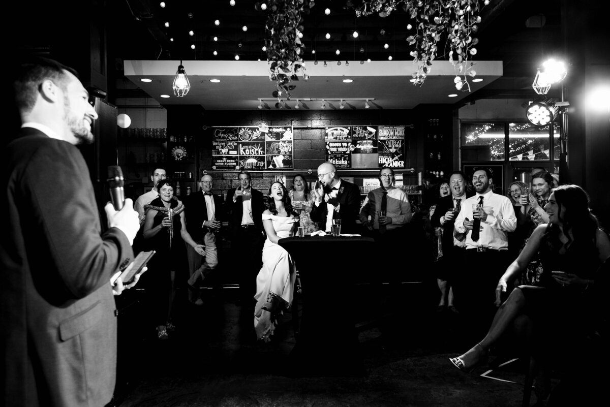 Black-and-white-photograph-of-the-best-man-giving-a-toast-in-the-foreground-and-the-bride-and-groom-at-their-sweetheart-table-in-the-center-of-the-reception-space-cracking-up-laughing-at-Eventide-Brewing