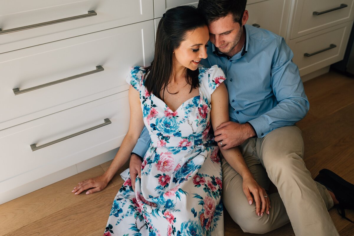 Man and wife sitting on floor in kitchen together, during bun in the oven pregnancy announcement photography session.
