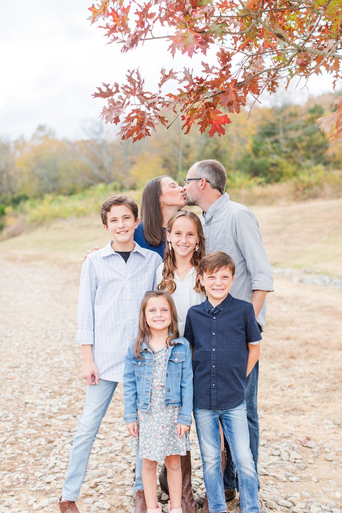 Husband and wife kissing while 4 kids smile at the camera underneath a pretty fall tree