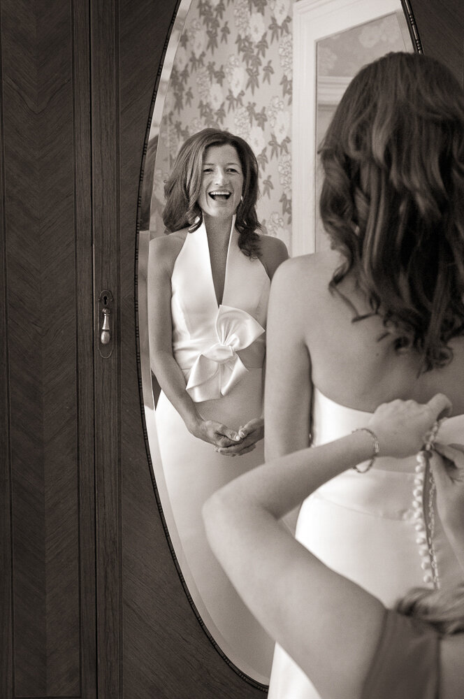 brunette bride with natural hairstyle laughing and looking in the mirror while having her v-neck, modern wedding dress buttoned at the back