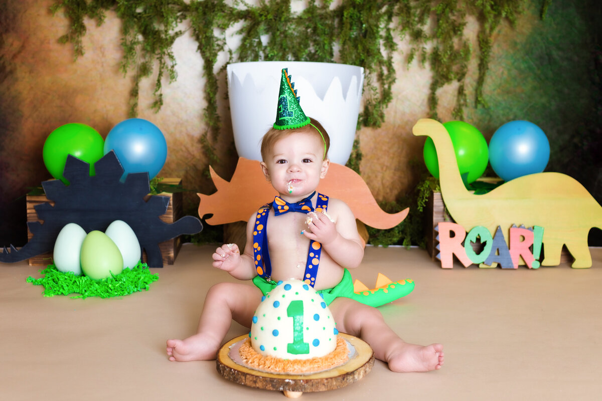 Cake Smash Photographer, a baby sits before a cake shaped as a dinosaur eggs, there are dinosaur cutouts behind him