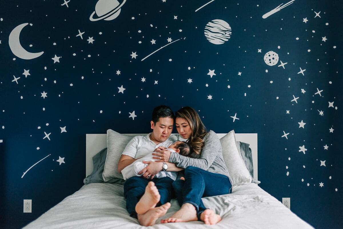 mom and dad snuggled in Alexandria home with newborn son against a navy space mural