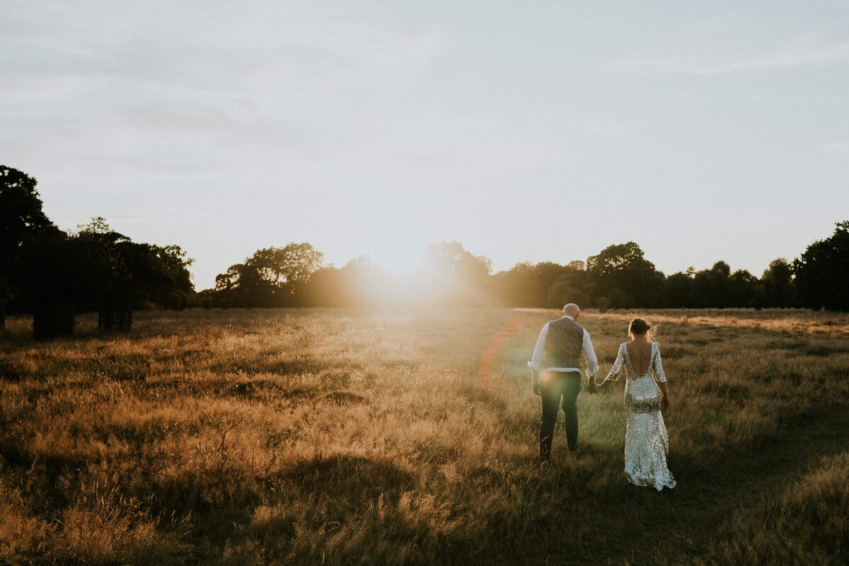 A bride and groom walking through a field at sunset