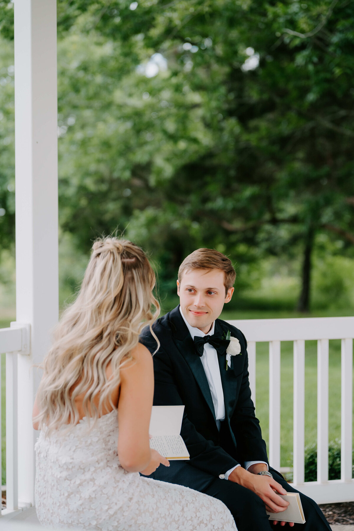 A couple sits on a porch, the woman with long blonde hair in a white gown, and the man in a black tuxedo. They exchange letters, surrounded by greenery.