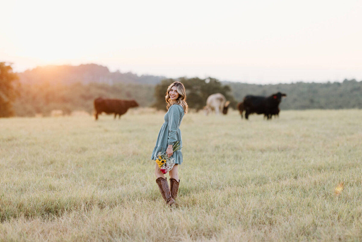 senior girl in teal dress and cowboy boots walking through open field at sunset with wildflowers in hand