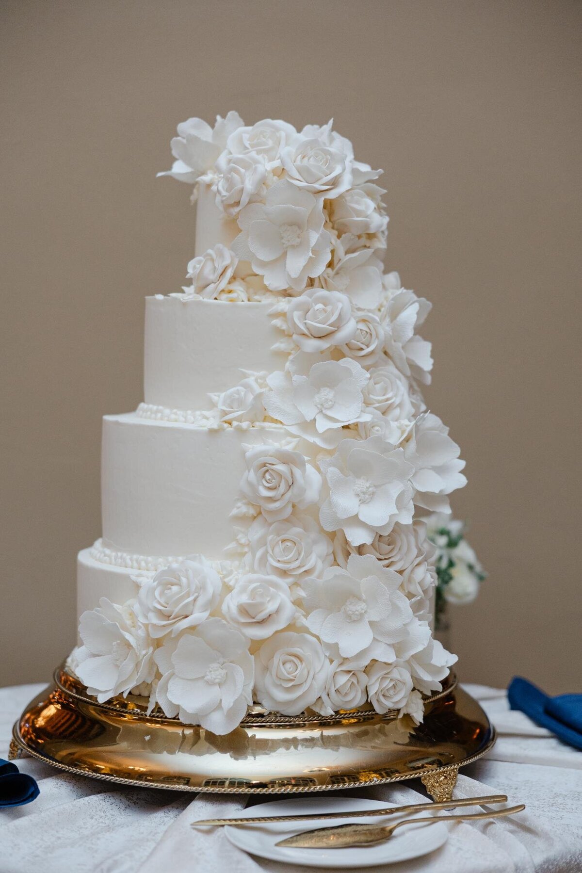 A multi-tiered white wedding cake adorned with delicate white flowers, displayed on a gold plate with serving utensils.