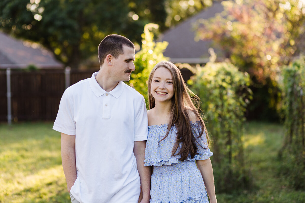 A couple holding hands in their backyard during their engagement session in DFW, Texas. The man on the left is looking at the woman and is wearing a white, short sleeve, collared shirt. The woman on the right is wearing a detailed, light blue dress. Lots of greenery can be seen behind them.