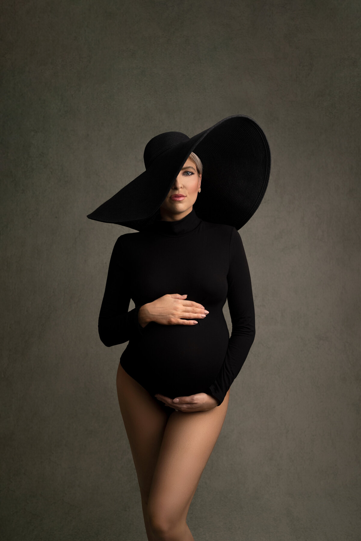 Woman poses for fine art maternity photos with New Jersey's best maternity photographer Katie Marshall. The woman is facing the camera wearing a black long-sleeve crew-neck bodysuit with bare legs. One hand is under her bump, the other hand is above her bump. She is wearing black hat with an oversized brim that partially covers the left side of her face.