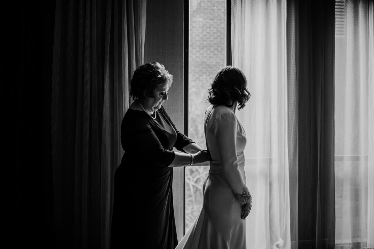 Baltimore wedding photographers captures a sentimental moment between the mother of the bride and the bride as the mother buttons her daughters gown and the bride stands in front of a window