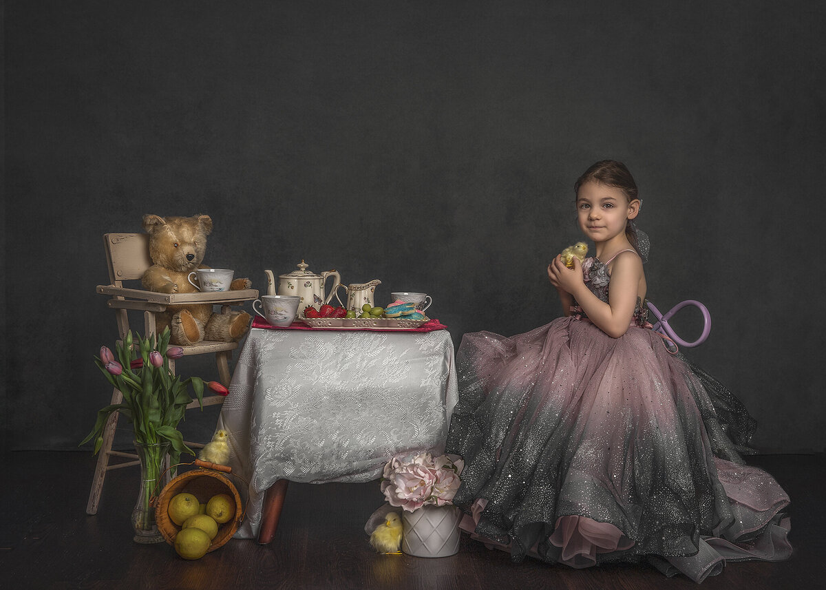 A young girl in a pink and gray gown sits across from her teddy bear at a picnic.