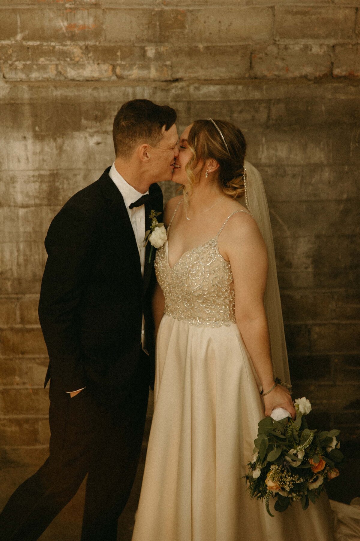 A bride and groom touching foreheads, smiling, in an intimate pose against a brick backdrop at an Iowa wedding, the bride holding a bouquet.