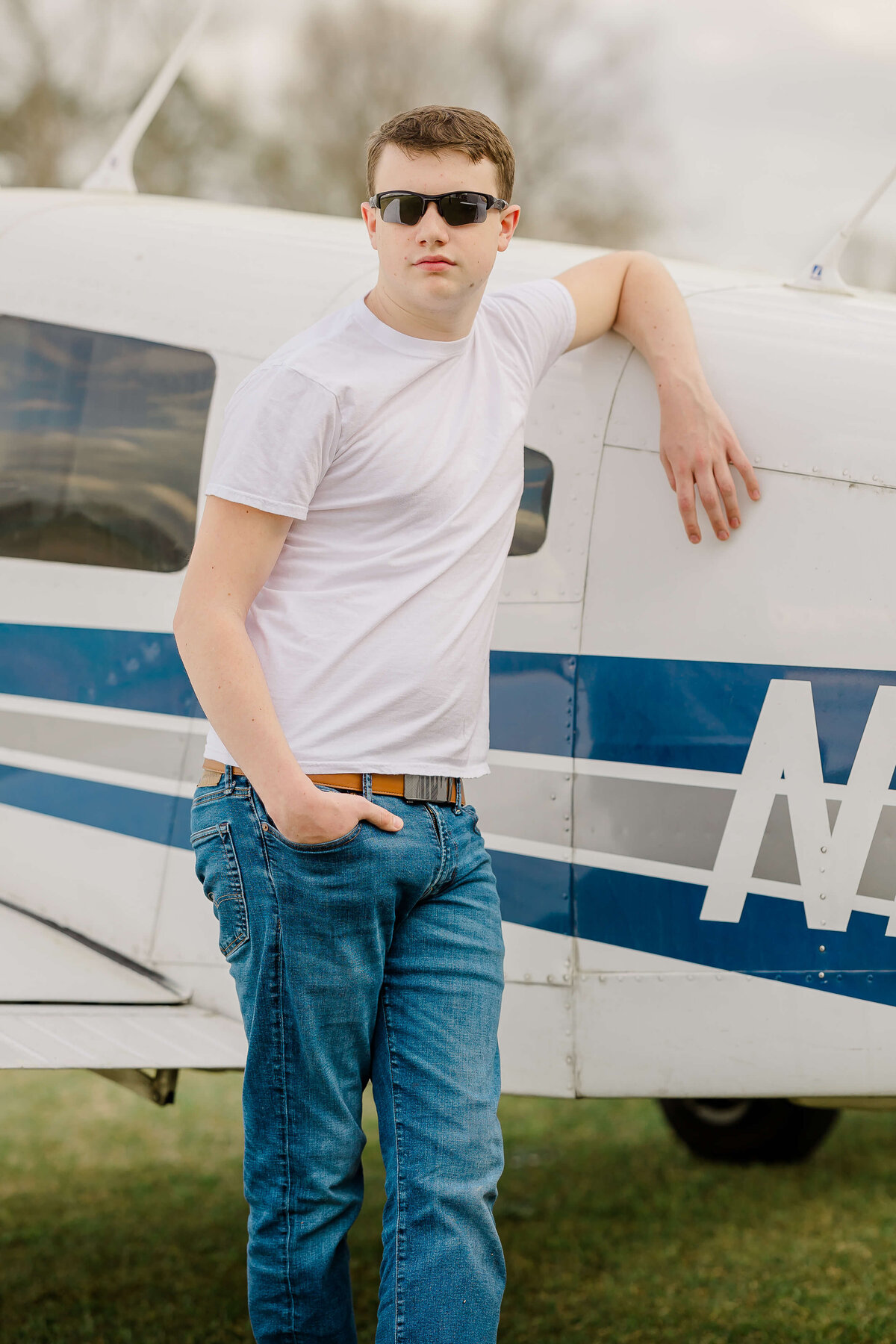 A person wearing sunglasses, a white t-shirt, and jeans leans against a small aircraft with blue and white markings. The setting appears to be an outdoor airfield with grass and a cloudy sky in the background.
