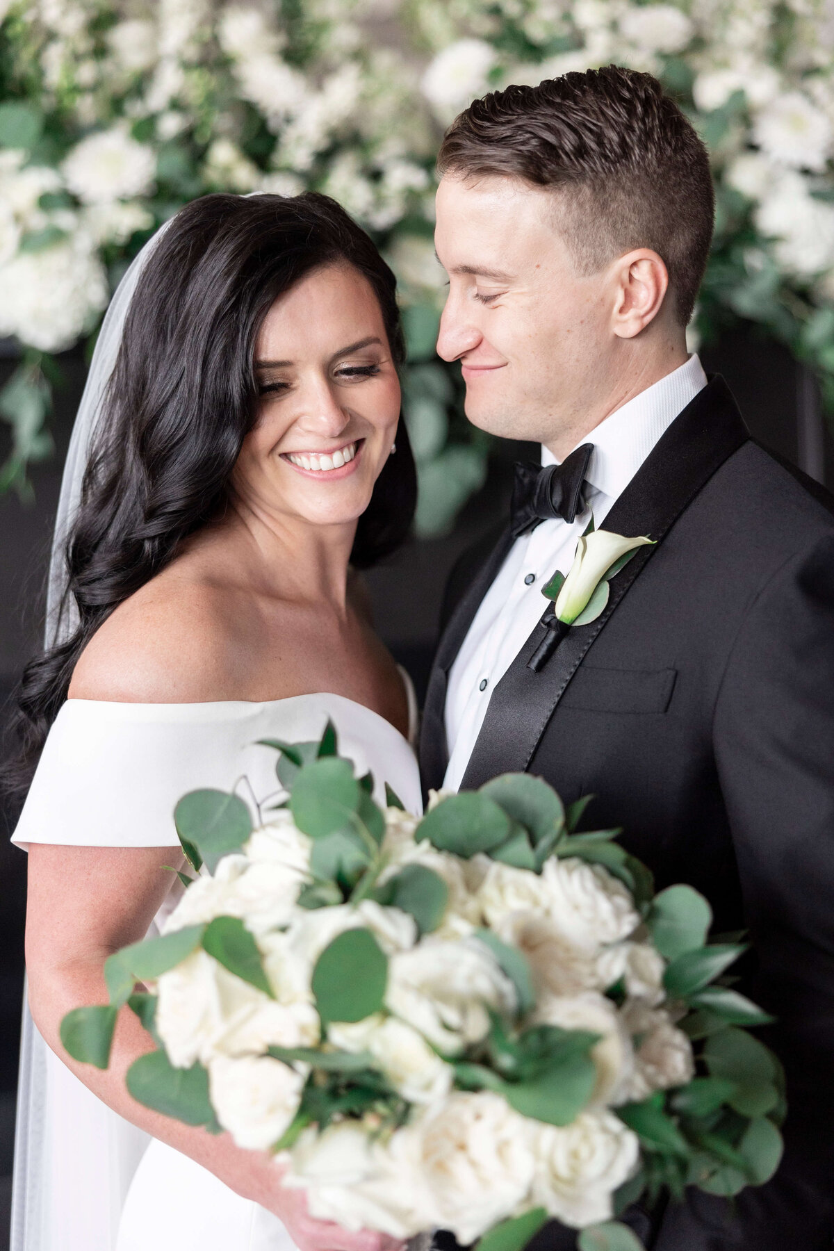 A bride and groom smile joyfully at each other while posing with a lush bouquet of white roses and greenery. The bride wears an off-shoulder wedding dress, and the groom is in a black tuxedo with a white calla lily boutonniere.