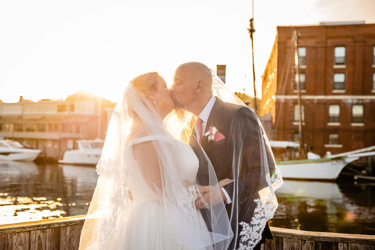 A bride and groom share a kiss on a dock, bathed in the golden light of the setting sun, with waterfront scenery behind them