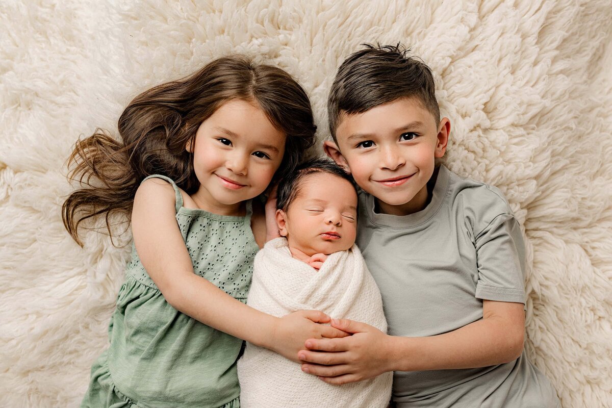 brother and sister dressed in green hold thier neborn baby brother between them while laying on a cream rug