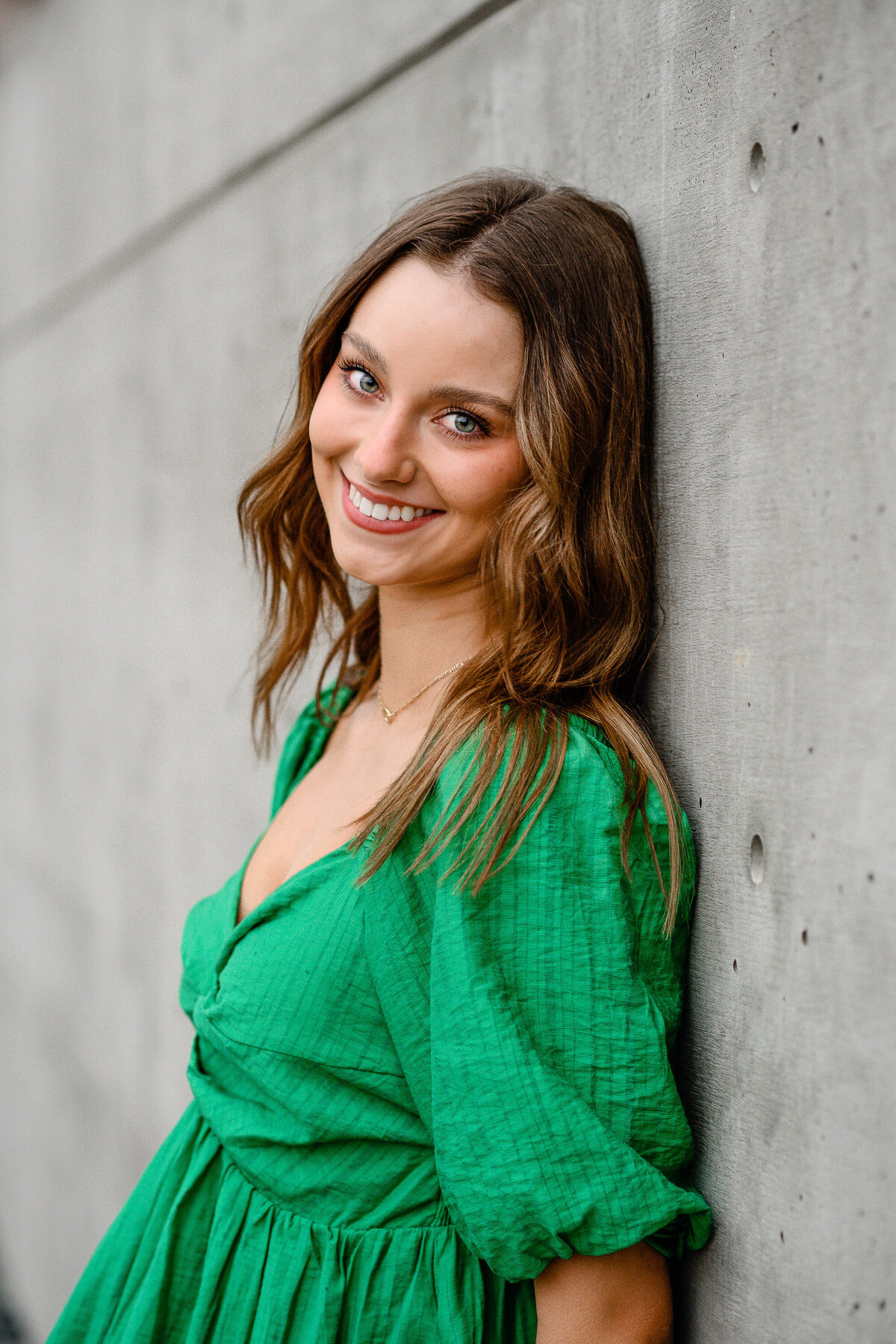 senior photo outfits with girl ina. green dress smiling over her shoulder as she leans against a concrete wall for her senior photos