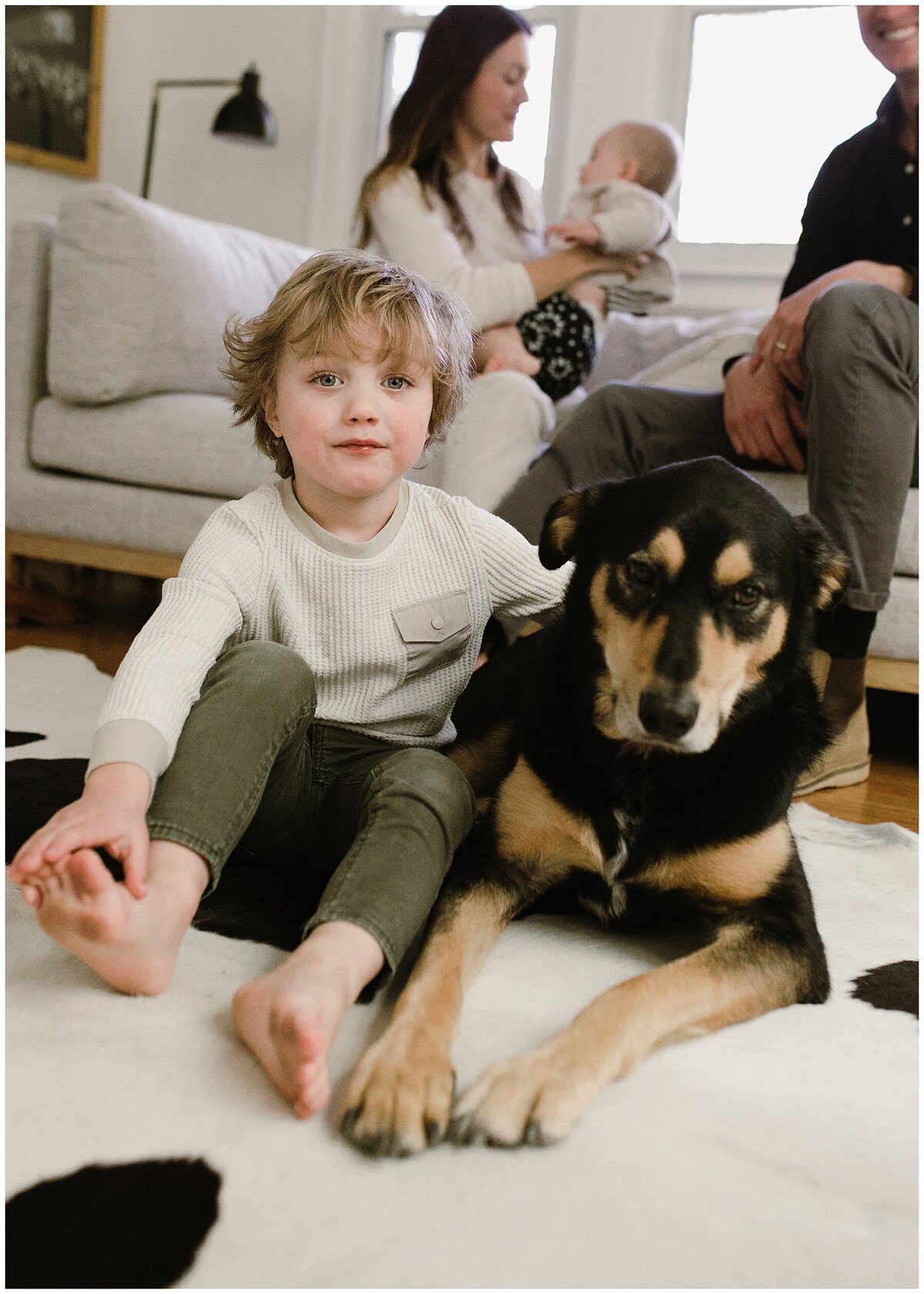 Child sitting on a rug next to dog with parents and baby in the background