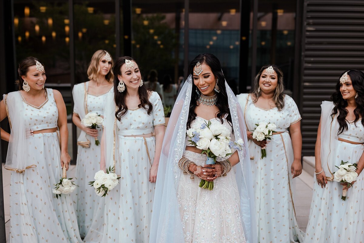 Hindu bridesmaids weairng light blue and gold sarees laugh with the Indian bride wearing a white beaded saree and light blue dupatta veil as she holds a large white bridal bouquet of hydrangeas for her Nashville wedding.