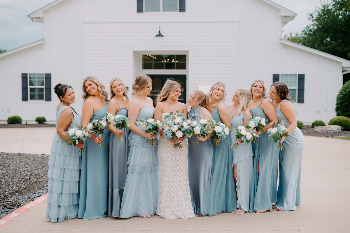 Bride in a white gown stands with bridesmaids in light blue dresses holding bouquets. They are smiling in front of a white building, conveying joy.
