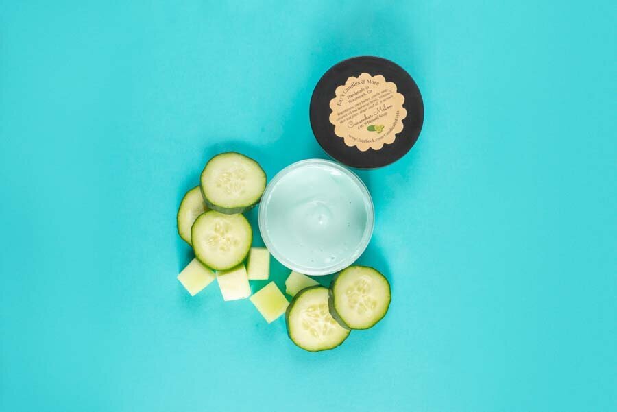 A skincare jar with a label next to sliced cucumber and shea butter cubes on a turquoise background.