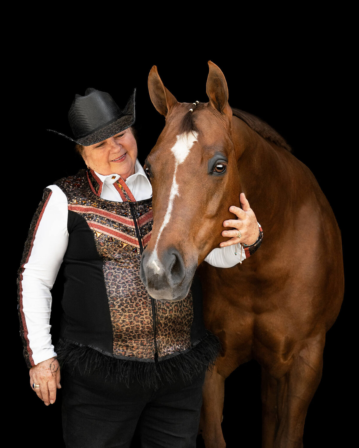 equestrian in leopard vest and black hate standing next to and looking at her Chestnut Quarter Horse Mare photographed against a black background.