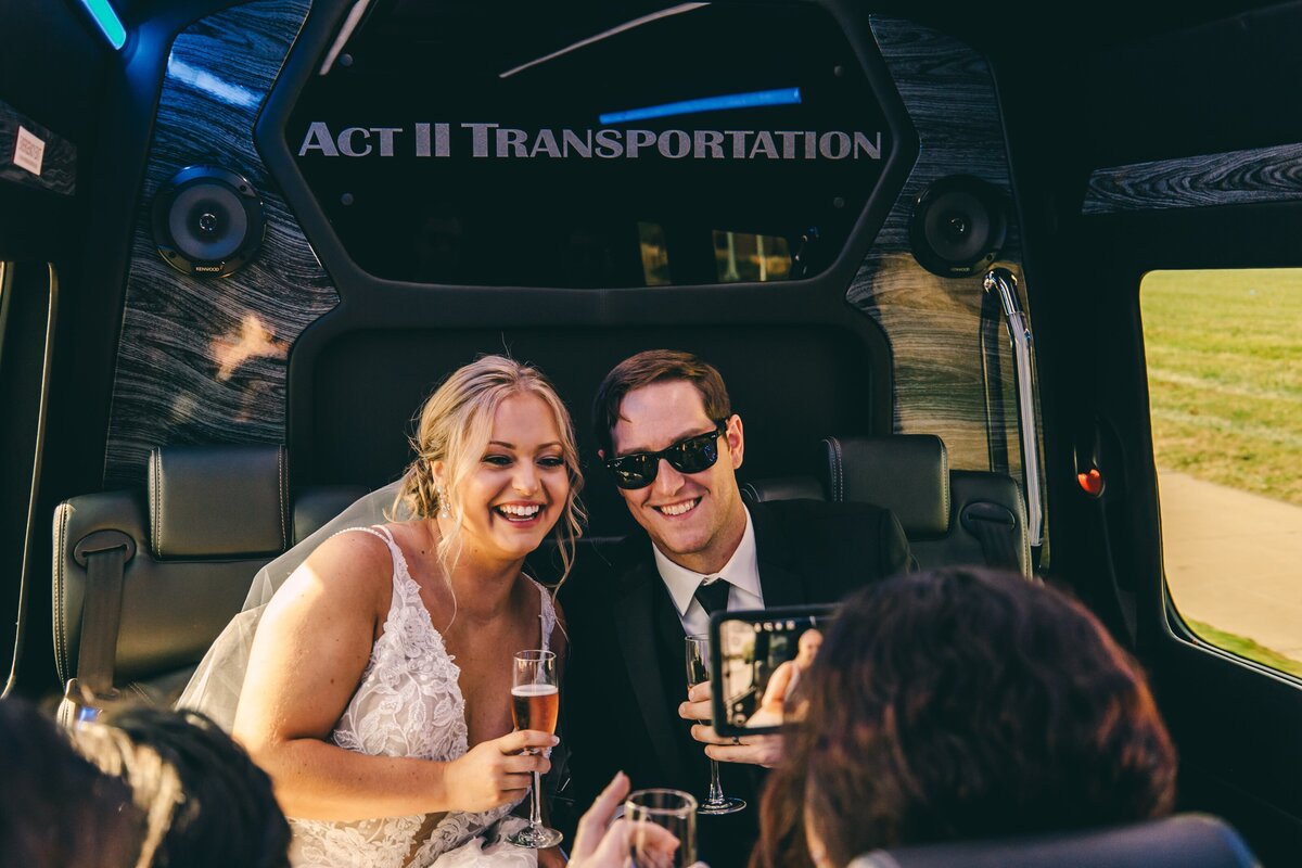 Bride and groom smiling inside a limousine, holding champagne glasses, while their wedding coordinator takes their photo with a smartphone.