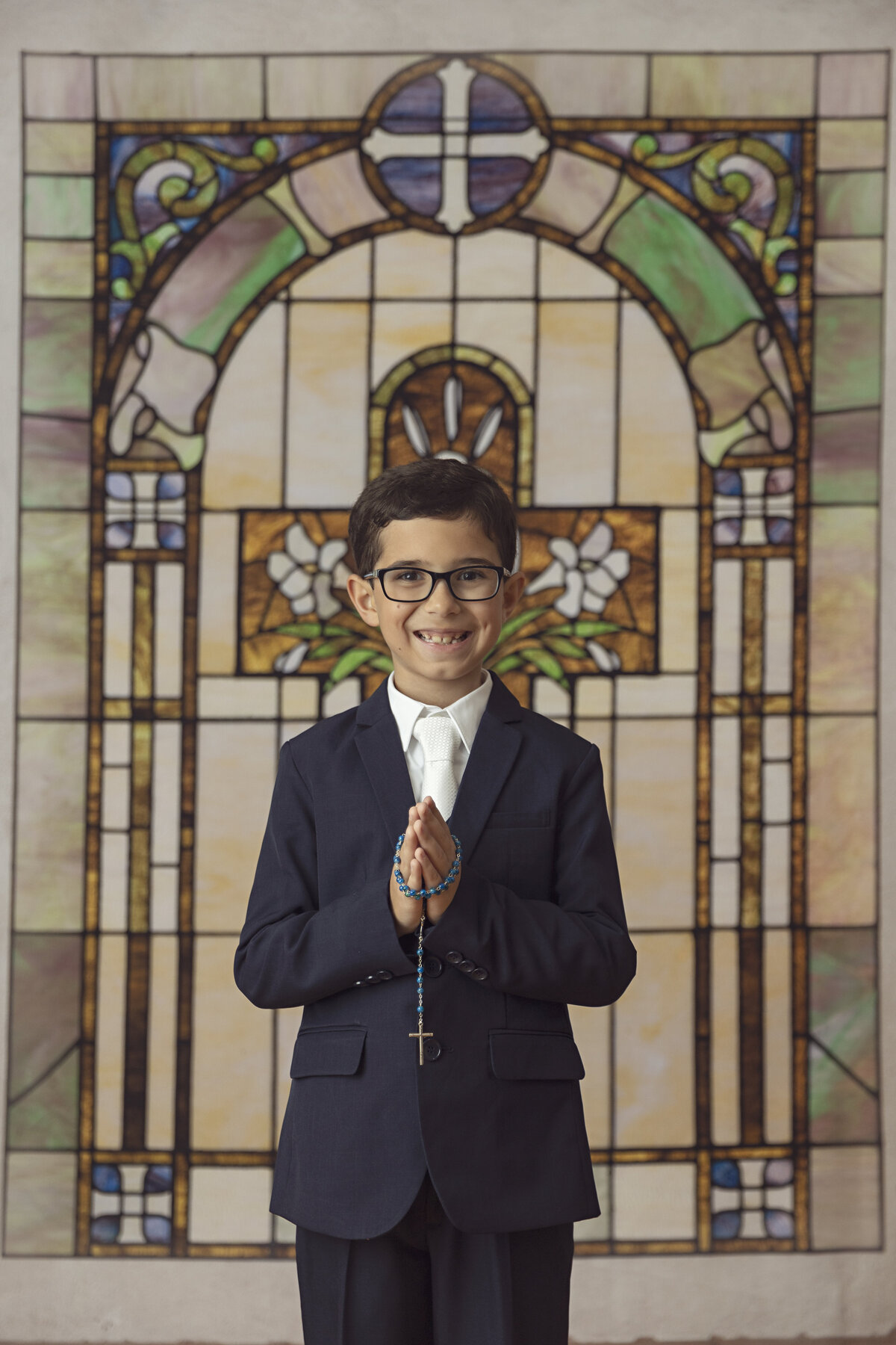 A young boy stands in front of a stained glass window praying with rosary beads in a black suit