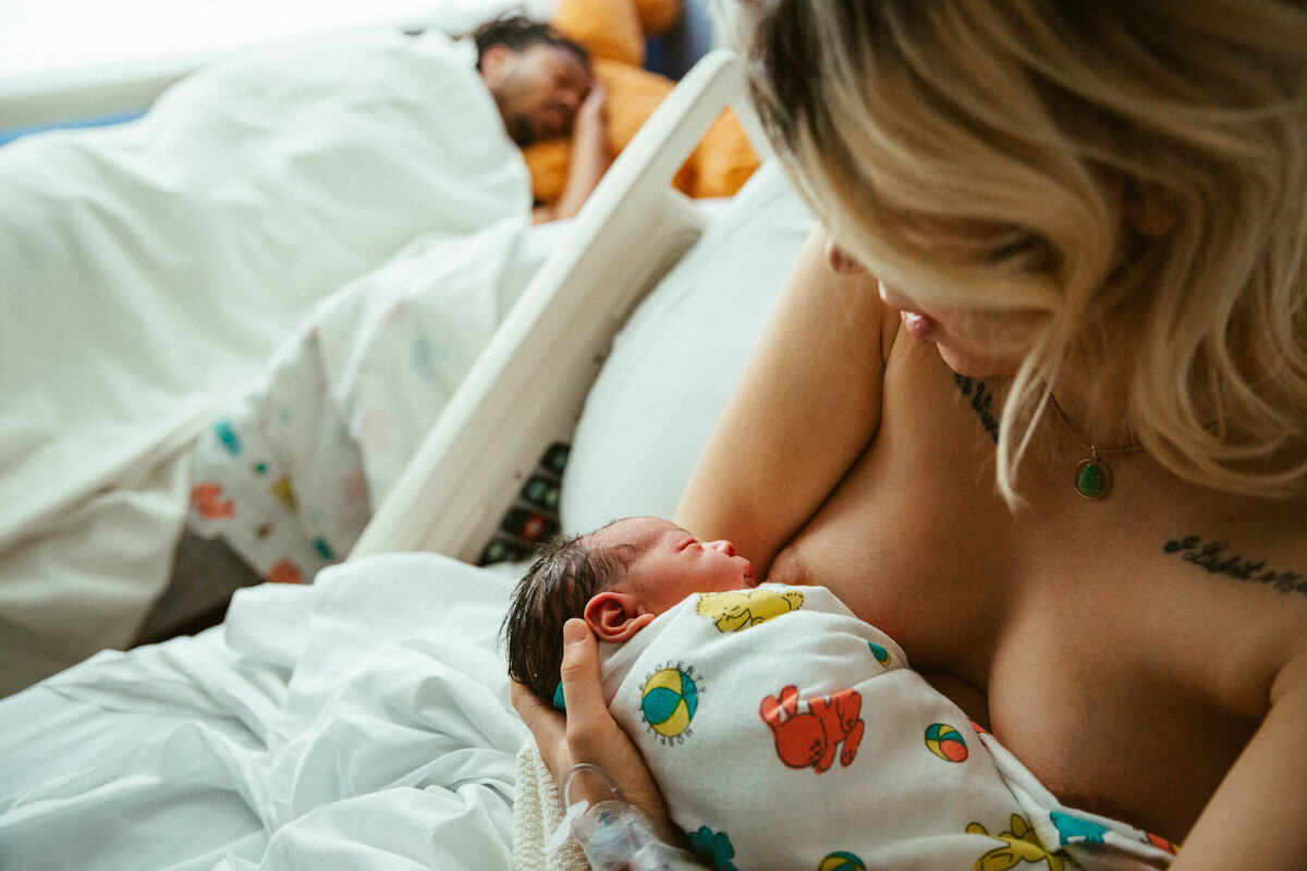 mom sitting in hospital bed nursing her newborn son while dad sleeps in the background