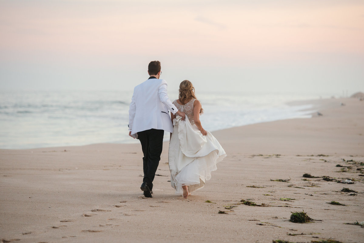 Groom holding his bride's dress as they walk on the beach at Oceanbleu Beachfront Catering