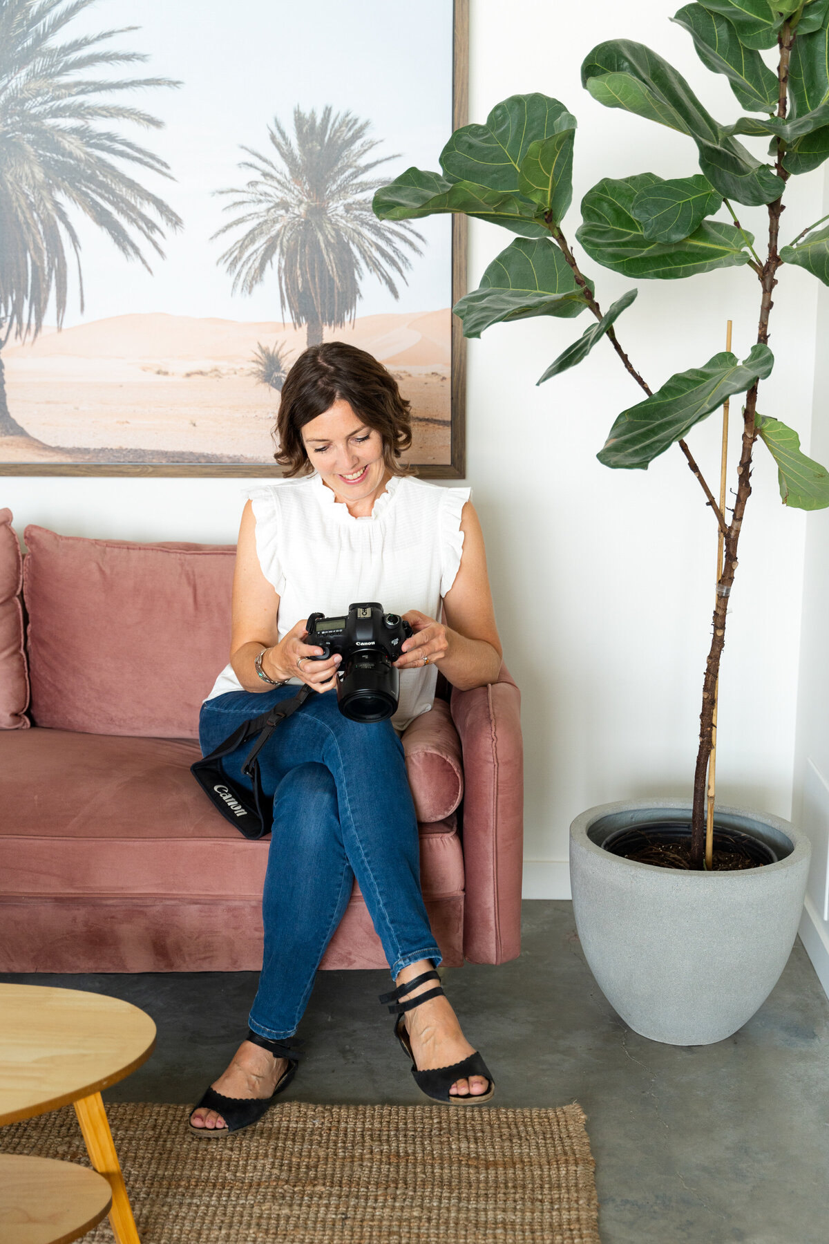 Calgary creative branding photographer, Kate Daniel Photography, sitting on a pink couch, wearing blue jeans and a white shirt, while looking down at her black camera.