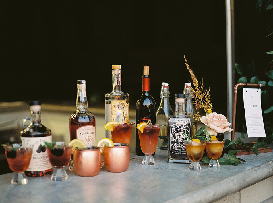 An assortment of cocktails amidst various alcohol glass bottles on a concrete counter with a drink menu and flowers.
