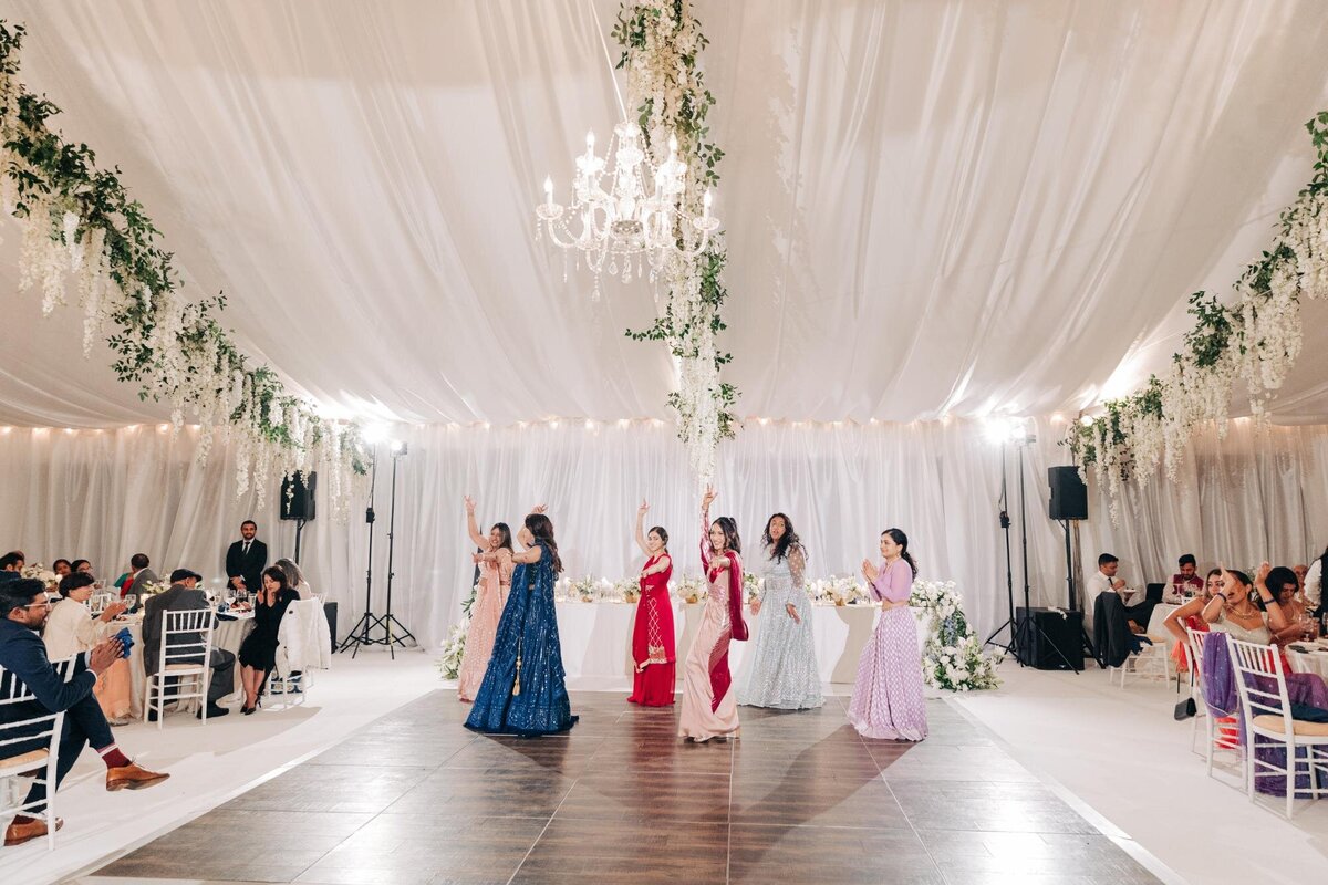 Elegant wedding reception in a decorated tent with guests seated and women in colorful dresses performing a dance.