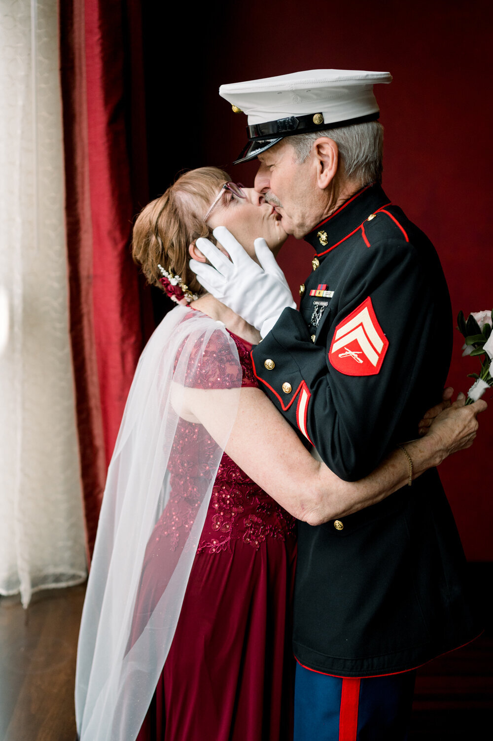 Marine in Dress Blues gently holding his wife’s face with white-gloved hands as they share a kiss before their 50th wedding anniversary celebration. She is dressed in a deep red floor-length gown with intricate beading and lace detailing, capturing a timeless and romantic moment.