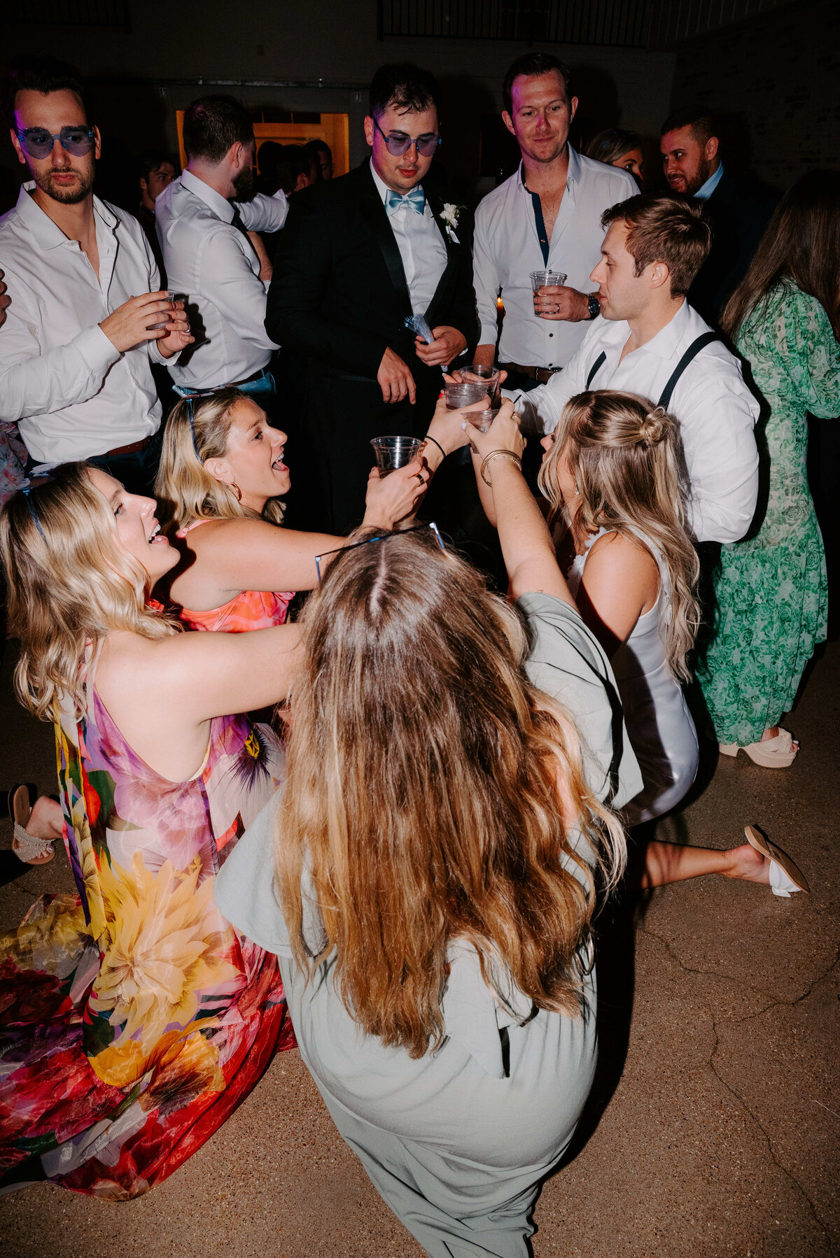 A joyful group of people in formal attire celebrate on a dance floor, raising their glasses in a toast. The scene is lively and filled with laughter.