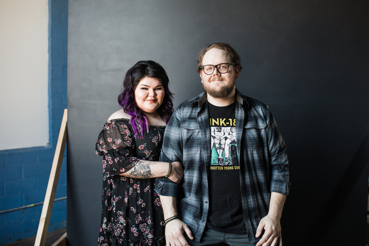 A couple smiling casually during their studio engagement session in Fort Worth, Texas. The woman on the left has her arms wrapped around the man's left arm. She has purple hair and is wearing a long black dress covered in flowers. The man on the right is wearing a black graphic t-shirt with a  blue flannel shirt on top, jeans, and glasses. They both are standing in front of a black background.