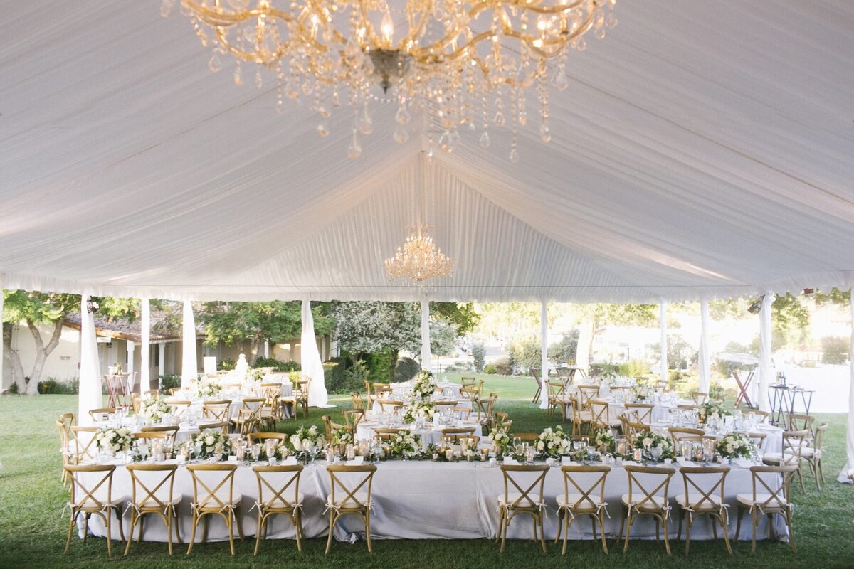 Tented-outdoor-wedding-reception-space, there are glass chandeliers above, white tbale cloths, and wooden chairs surrounding the round tables.