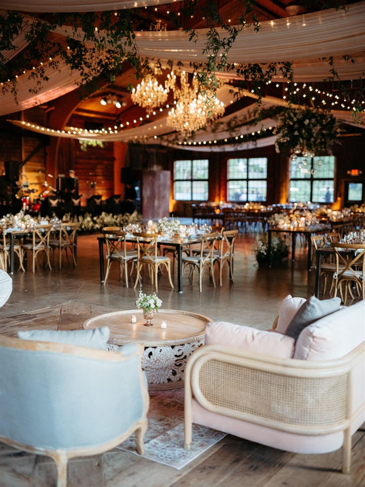Cedar Lakes Estate wedding venue reception lounge space in front of romantic tables, natural wood crossback chairs, candle-lit tablescapes, under chandeliers, twinkle lights, draped white fabric, and botanical swags.