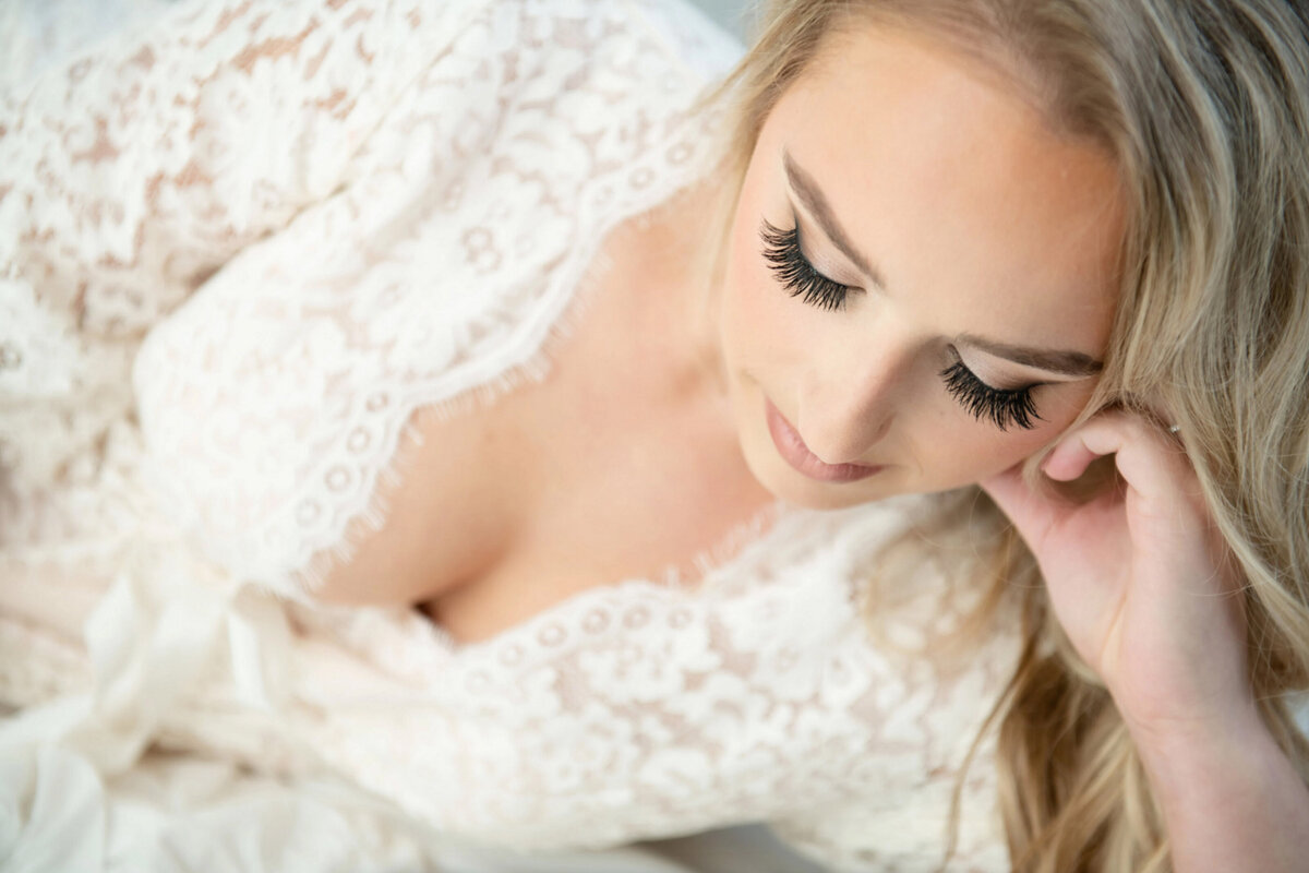 Bride posing in a white lace robe with her eyes down and long lashes